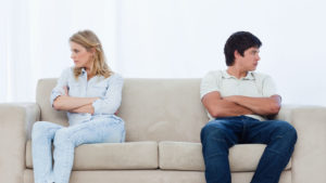 A angry couple sit at the two ends of the couch with their arms folded looking away