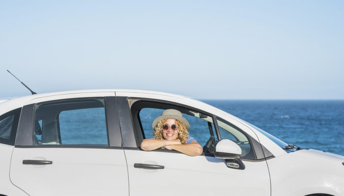 one beautiful happy woman looking at the camera from inside of a car with the sea or ocean at the background - adult traveler and nomad lifestyle and concept - outdoor sunny day